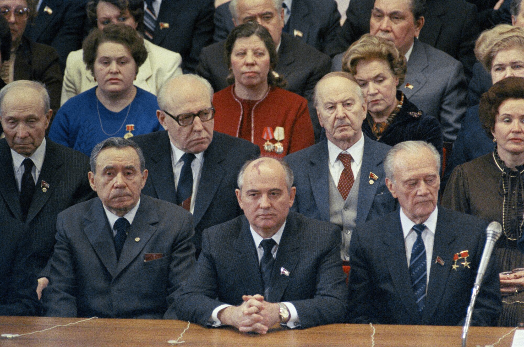 A man in suit and tie, hands clasped while seated, is flanked by other solemn-looking men and women 