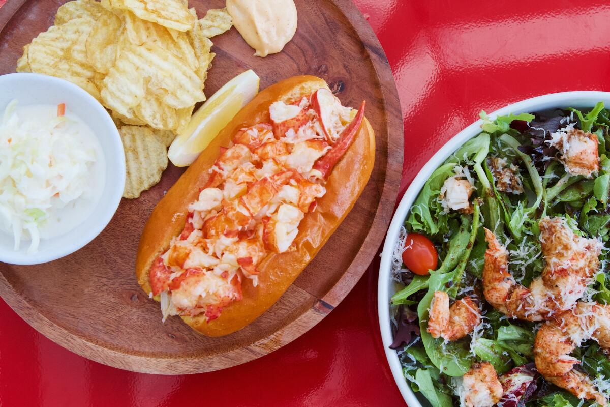 An overhead photo of a lobster roll and a lobster-topped salad on a red tabletop.