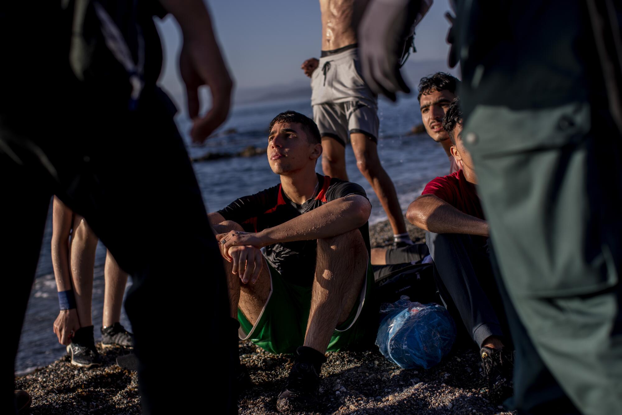 Teenagers and young men sit on the beach, still wet from their swim crossing, surrounded by Spanish guards