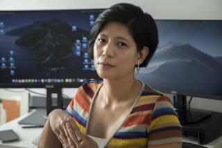 A woman in a striped shirt interlaces her fingers while sitting at a desk with two large computer monitors.