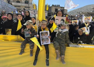 Children, some holding up photos of people, are flanked by adults