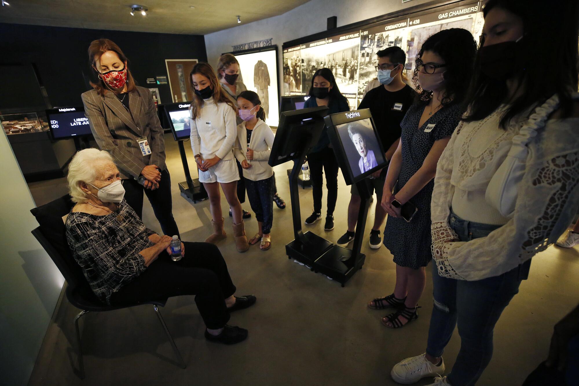 Students wearing face coverings listen with heads bowed respectfully as a seated woman speaks