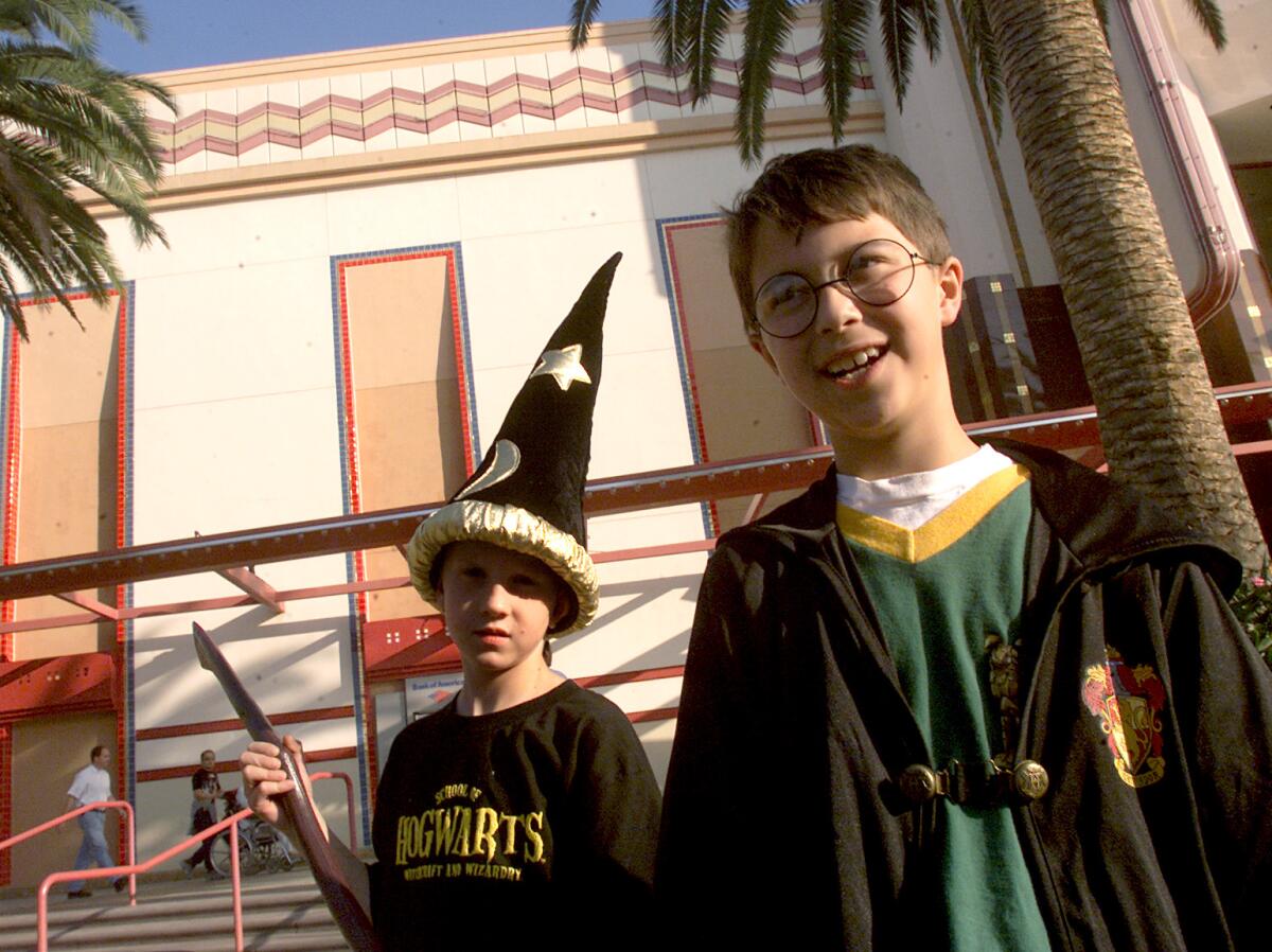 Two young boys in costume, one in a pointed hat and another in a robe, stand next to a movie theater and palm trees.