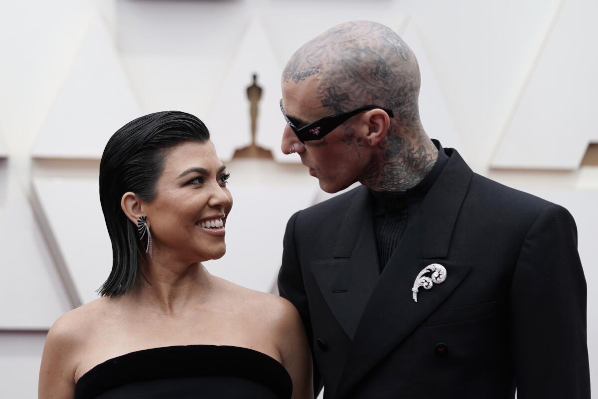 Kourtney Kardashian smiles at Travis Barker; both wear black formal attire while posing for photos in front of a white wall