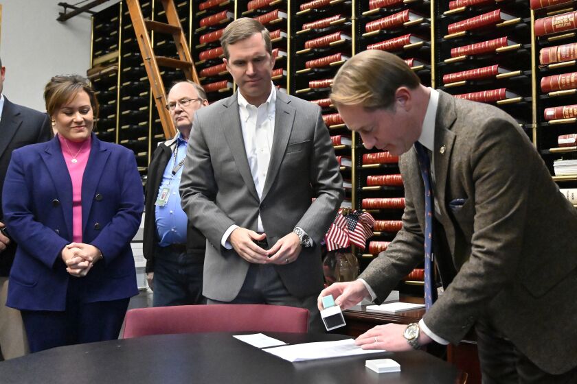 Kentucky Governor Andy Beshear, center, and Lt. Governor Jacqueline Coleman, left, look on as Secretary of State Michael Adams stamps the paperwork officially entering the race for reelection in Frankfort, Ky., Monday, Dec. 5, 2022. Fresh off attending an event trumpeting Kentucky's largest-ever economic development project, Democratic Gov. Beshear filed for reelection Monday. (AP Photo/Timothy D. Easley)