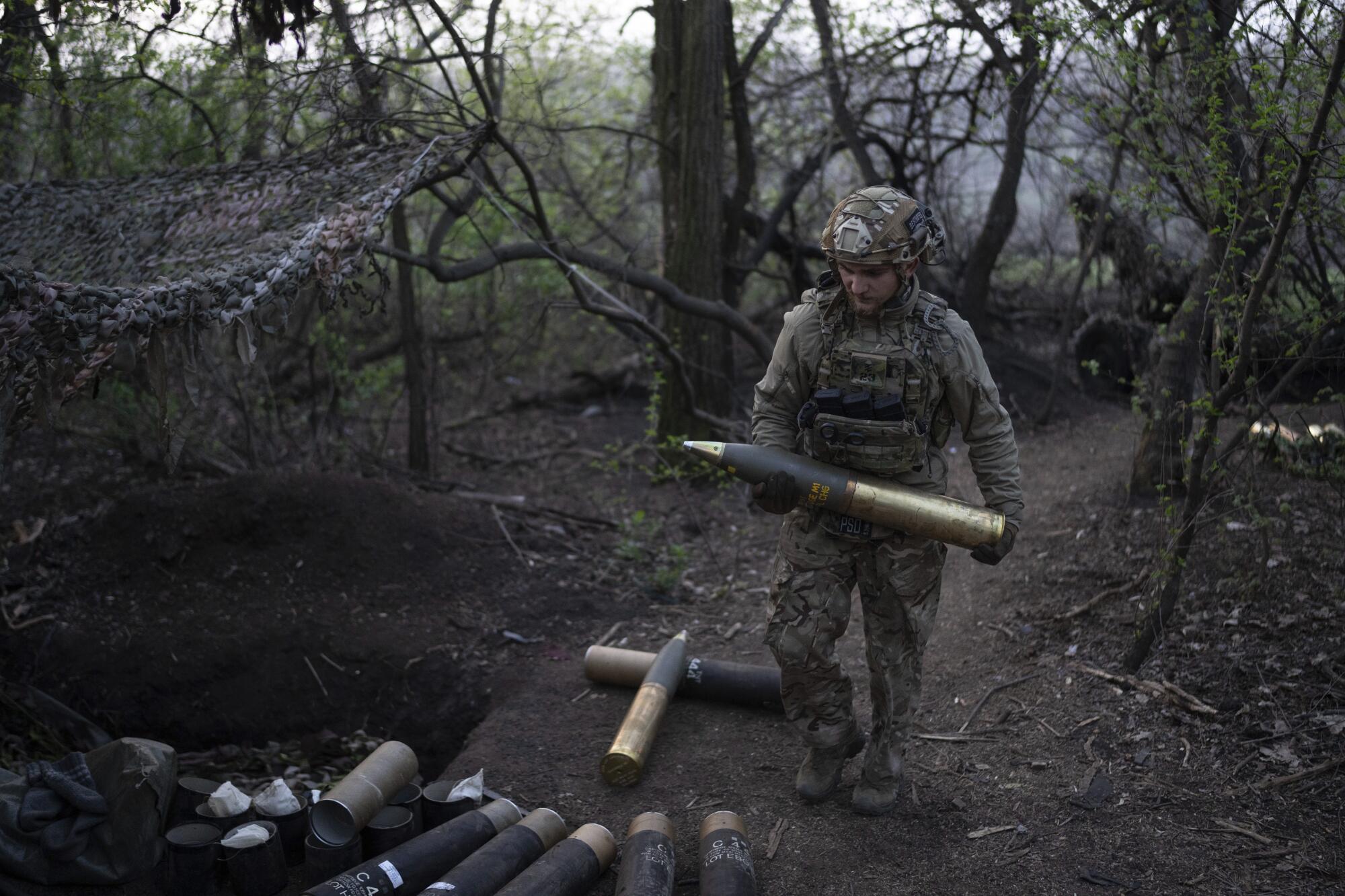 A person in camouflage, body armor and a helmet carries a large howitzer shell in a wooded area.