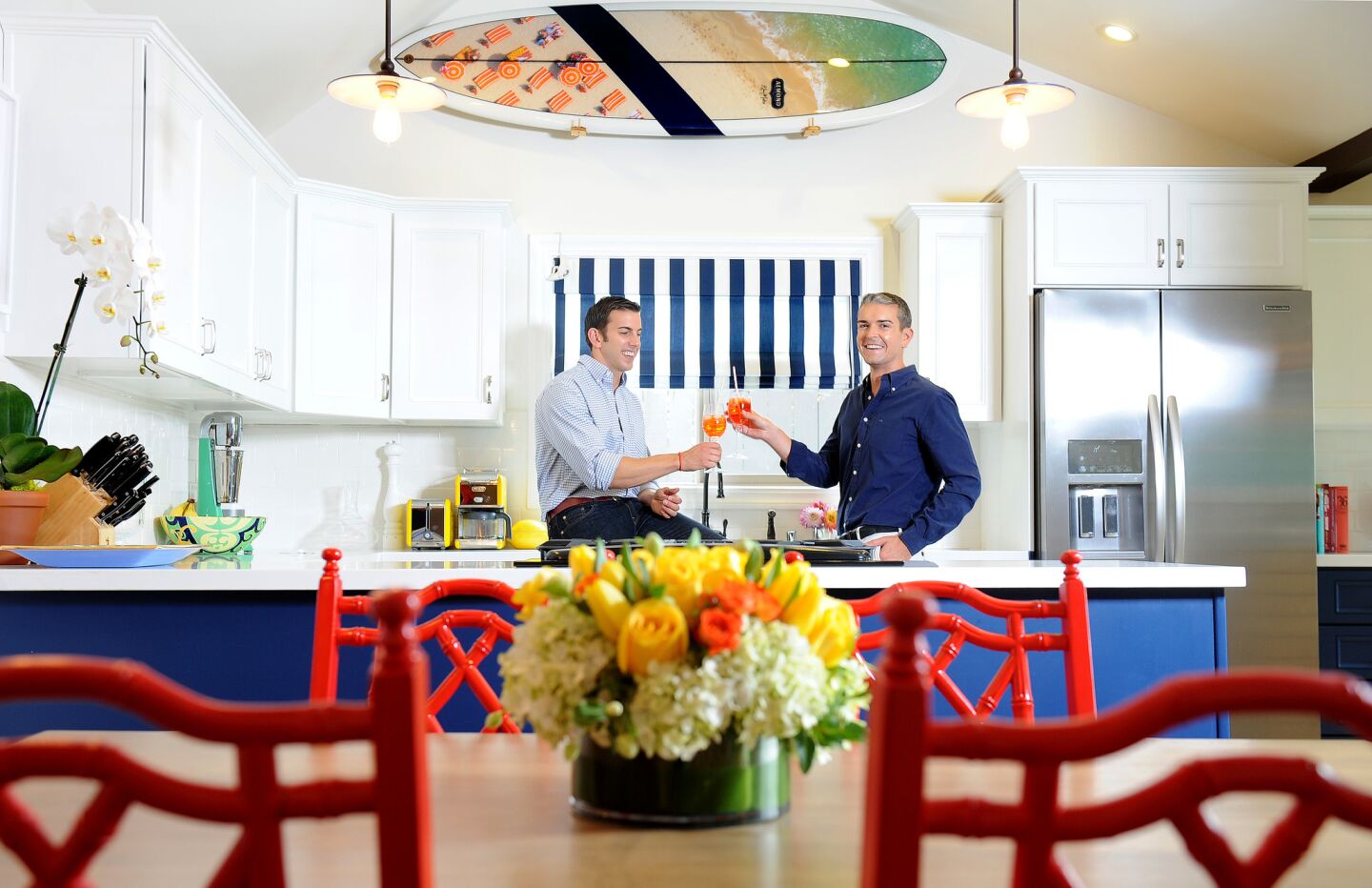 Jeff Richardson, left, and photographer husband Gray Malin share a toast in their kitchen. This part of the house has a preppy, nautical vibe, with white tile and navy blue cabinetry. The 1960s dining chairs, painted cherry red, feel almost Chinese.