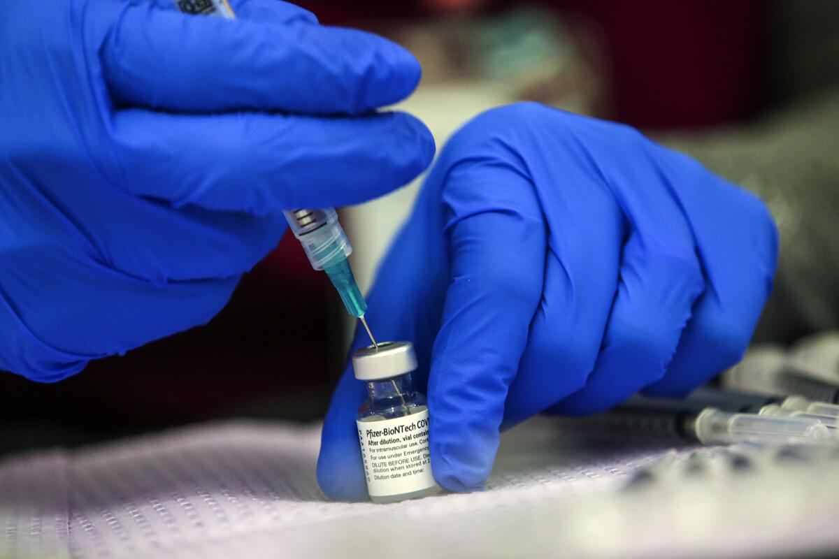 A gloved worker holds a Pfizer COVID-19 vaccine vial with one hand while another hand holds a syringe inserted in it.