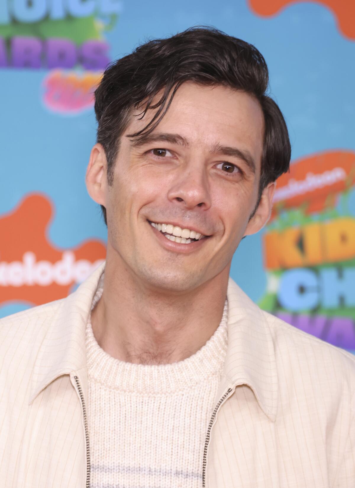 A man with short, wavy dark hair wearing a cream-colored sweater and jacket smiling against a background of many colors