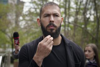 Andrew Tate holds the bottom of his beard while wearing a black t-shirt and blazer