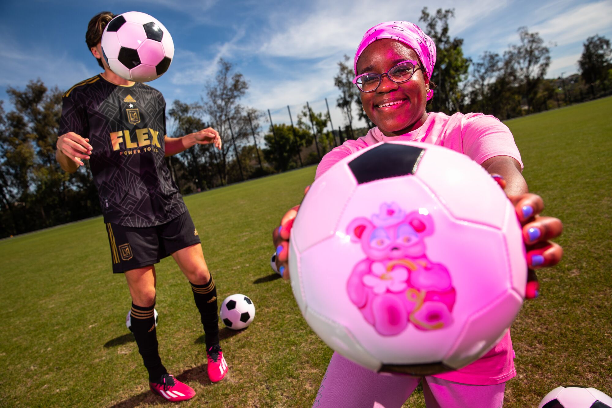 A woman in pink shows off a soccer ball, with a cute image,  as a soccer ball obscures a player's face in the background.