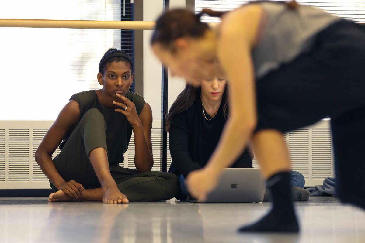 Two people wearing black sit on the floor, once looking at a laptop, the other watching a dancer moving in front of them.