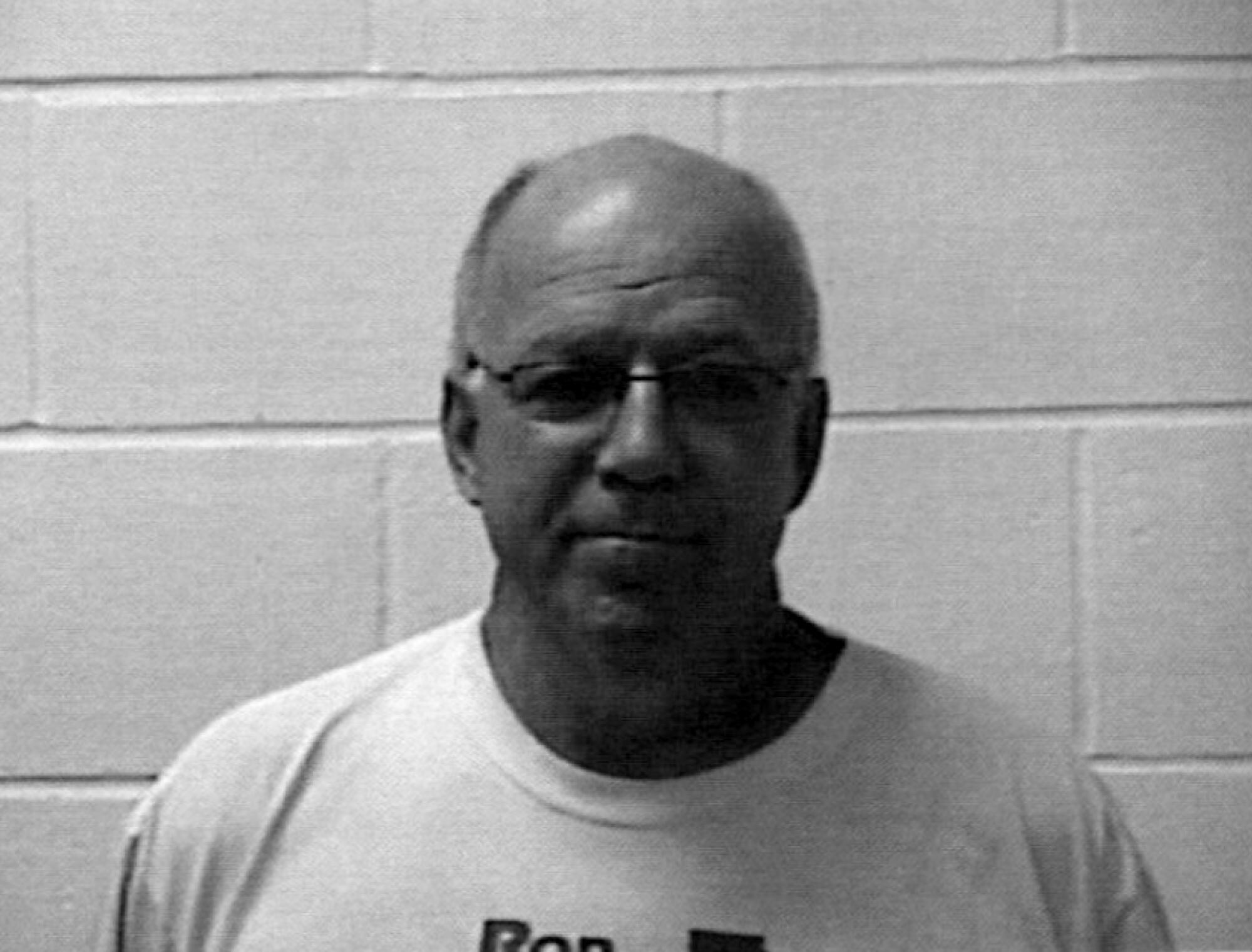 A black-and-white photo of Steve Kruse standing near a brick wall, wearing a T-shirt and glasses