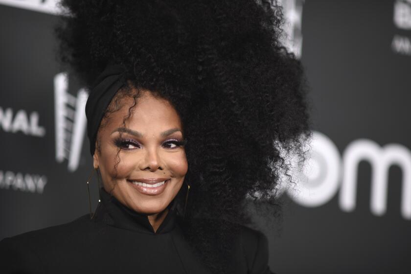 Janet Jackson wearing a black, high-neck top as she poses and smiles against a black background at a red carpet event