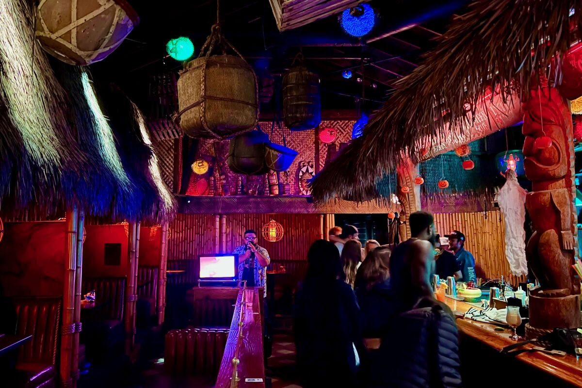 A man sings karaoke in a tiki bar, with patrons lined up at the bar