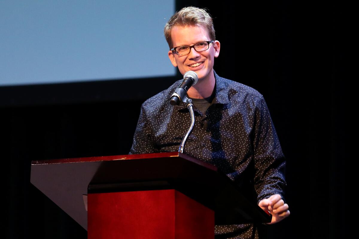 Hank Green, wearing rectangular glasses and speaking from behind a wooden lectern.