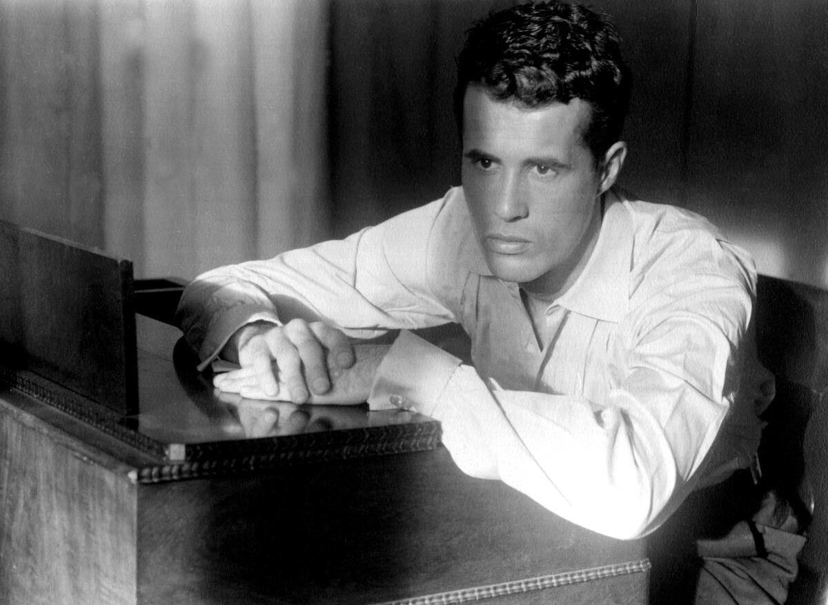 In a vintage photograph, Kenneth Anger is shown setead at a pianon in a white, button down shirt.