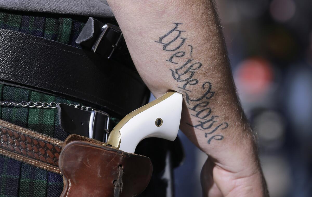 Closeup of the handle of a pistol and a man with a tattoo on his arm that says "We the people."