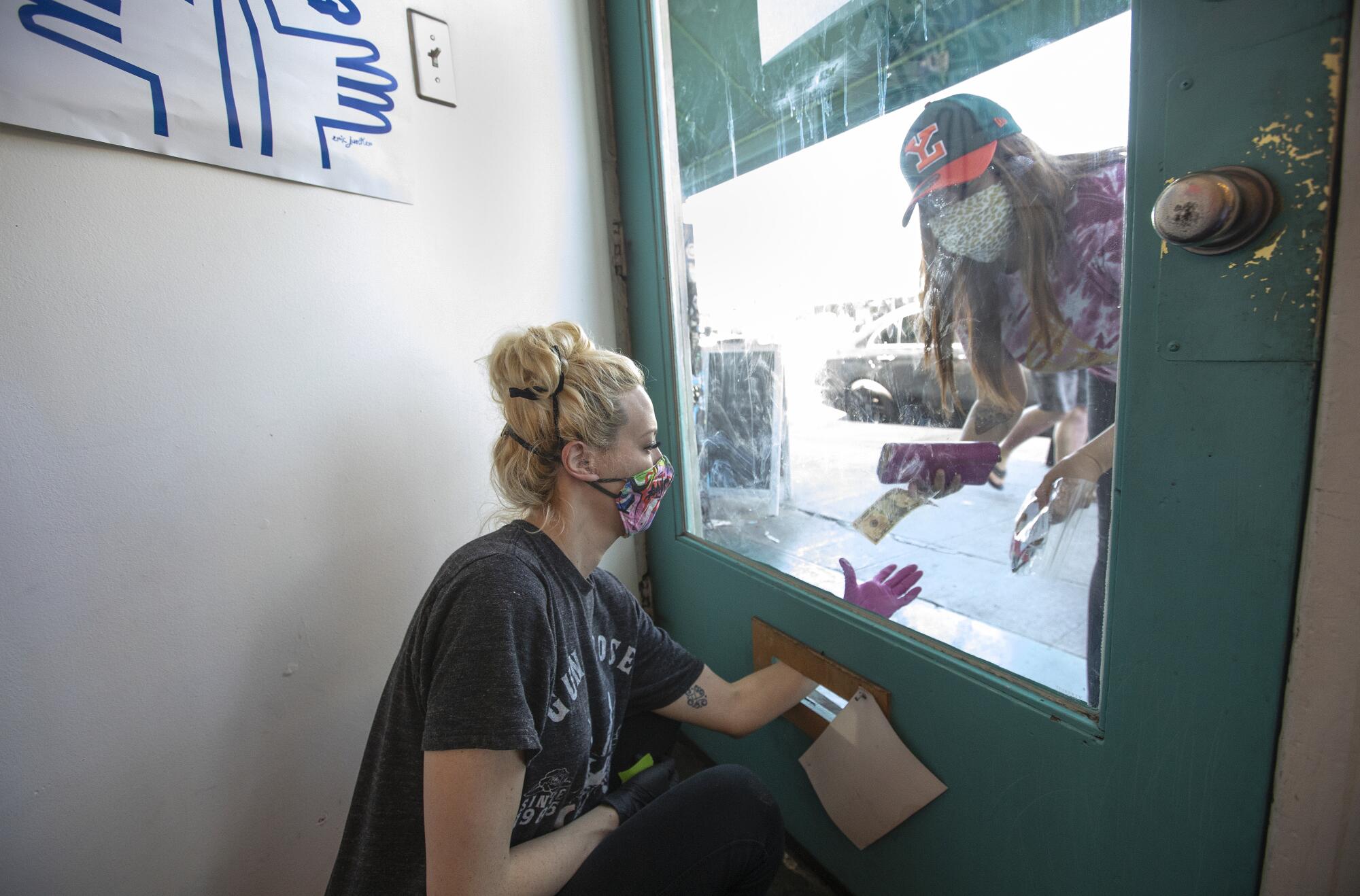 Christine Flick uses the mail slot to exchange money for a mask with a walk-up customer at the front door to Matrushka Construction in Silver Lake.