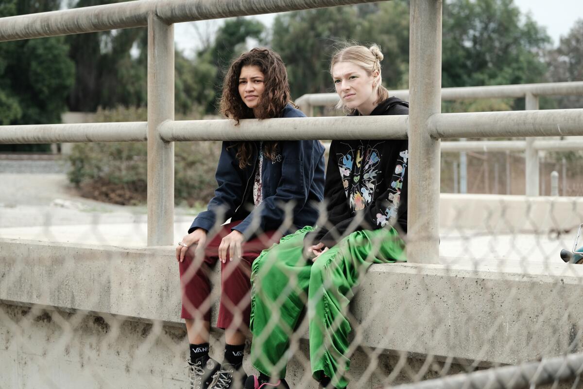 Zendaya and Hunter Schafer sitting on a slab of concrete behind a chainlink fence