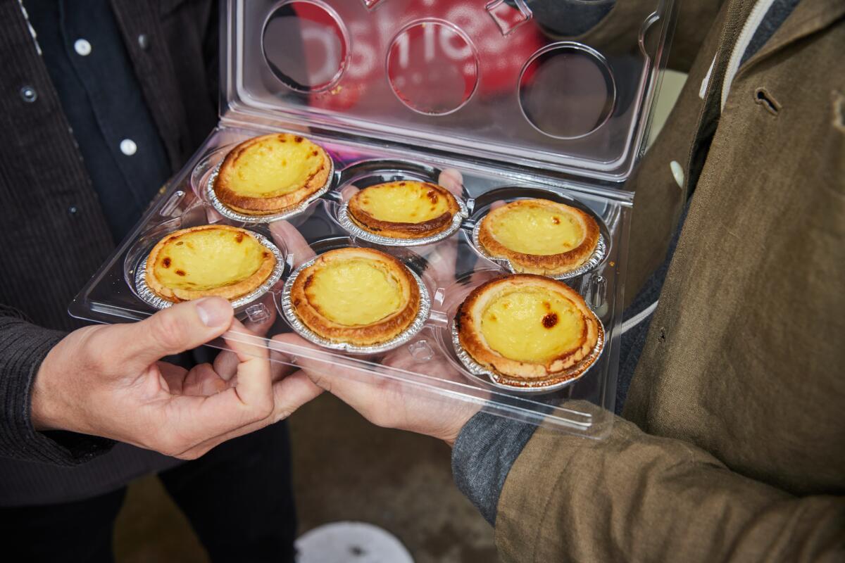 Hands hold a plastic container of six egg tarts.