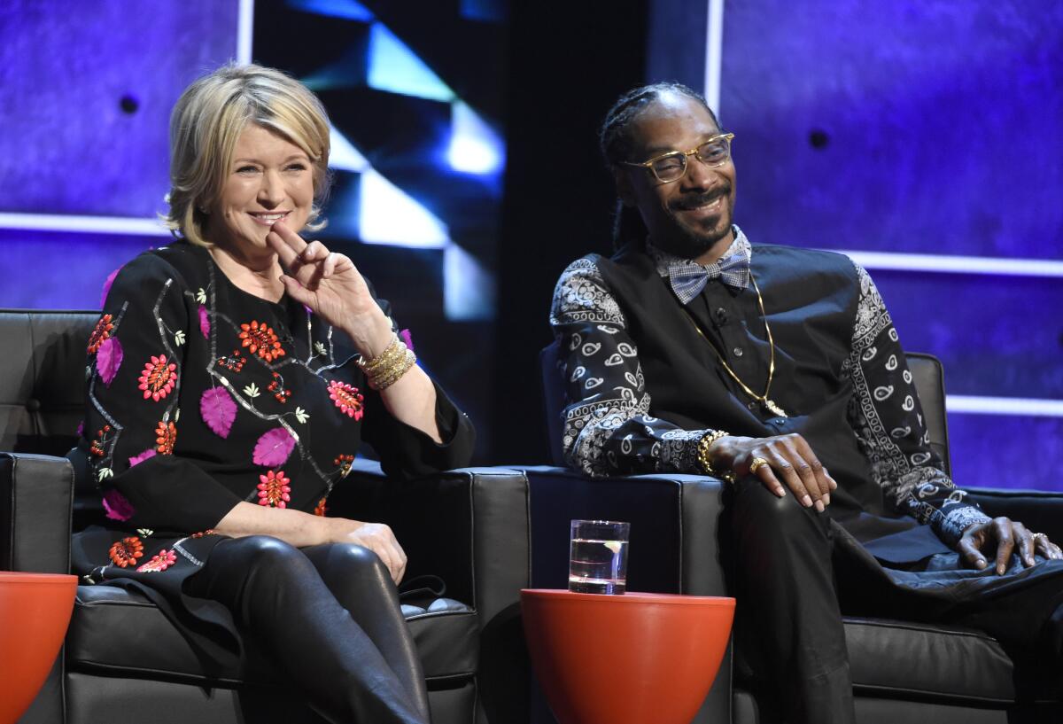 Martha Stewart and Snoop Dogg smile while sitting in leather armchairs on a TV set