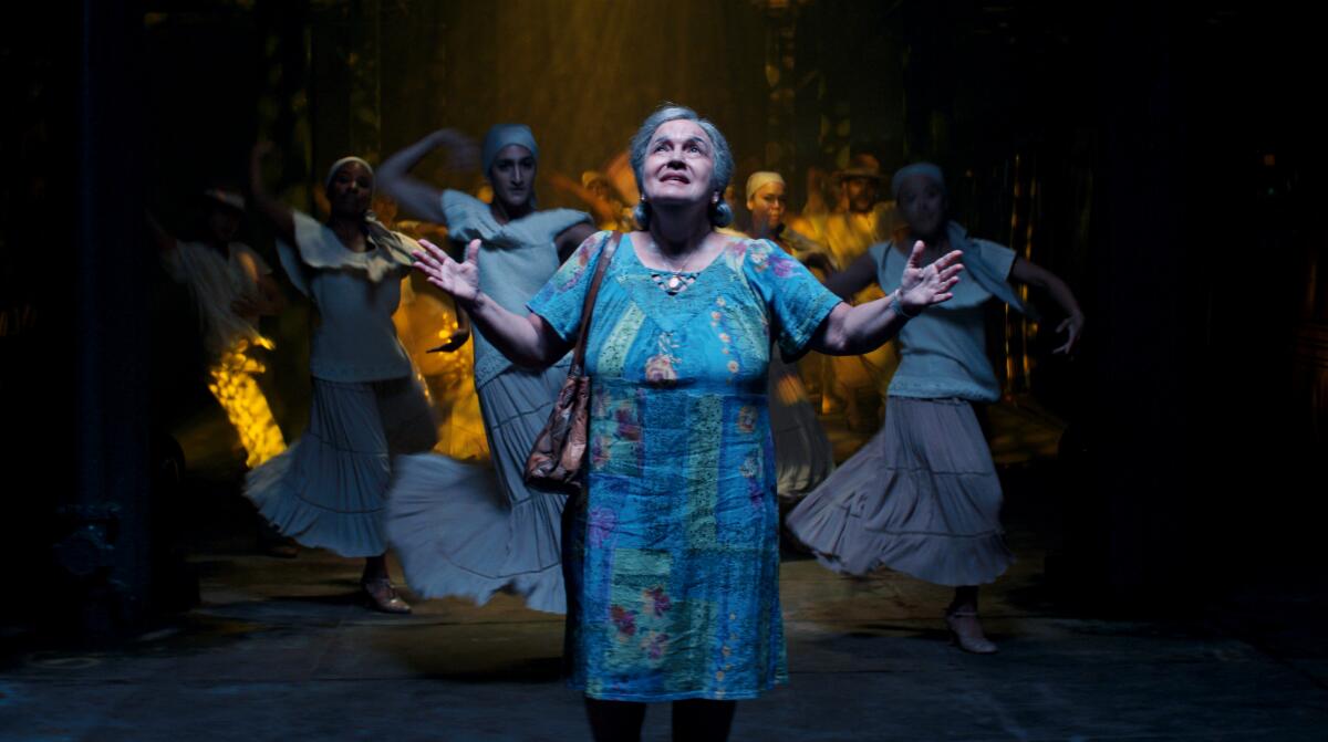 A woman looks up and spreads her arms as women dance behind her.