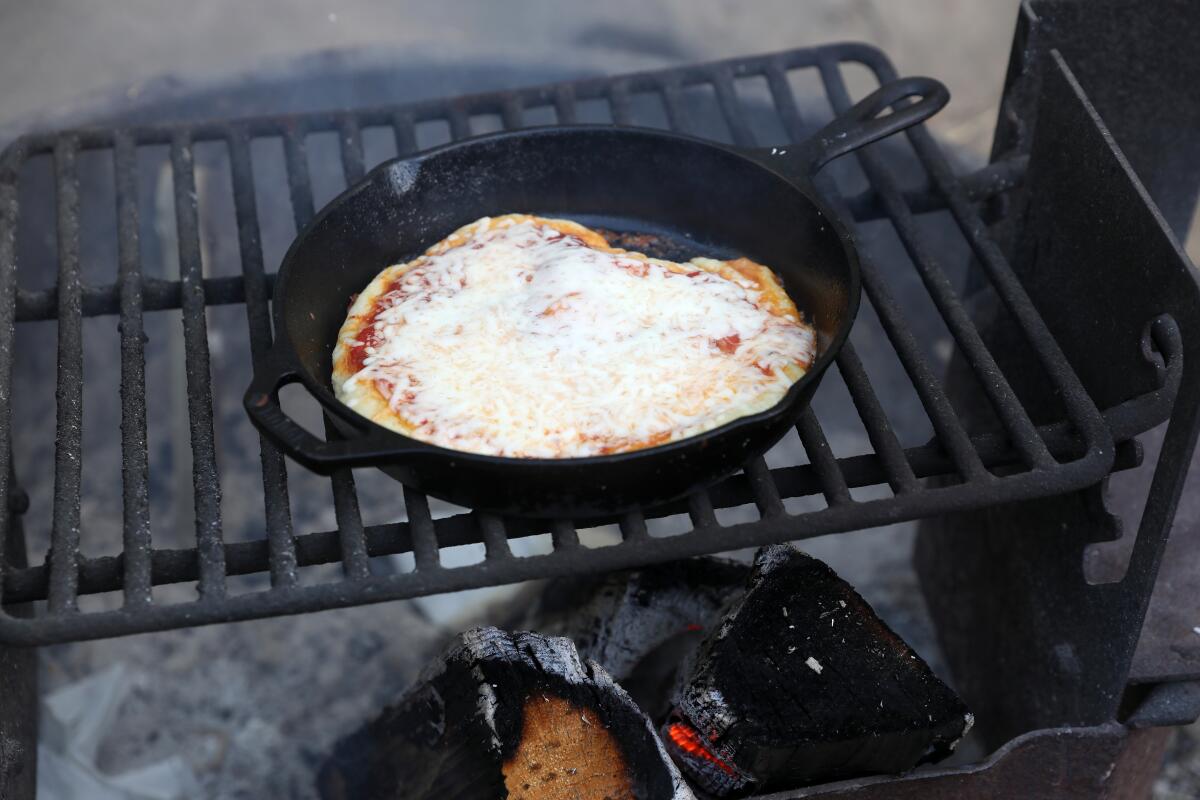 Pizza cooks over a campfire in a cast-iron skillet.