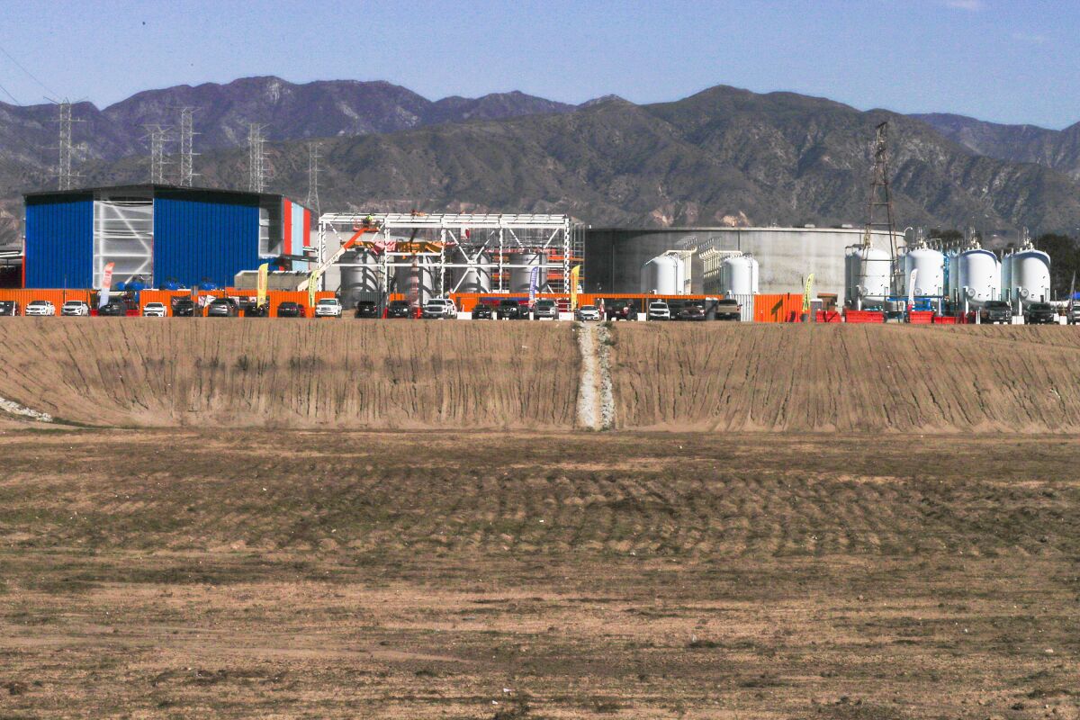 A group of closed aboveground tanks and buildings on land outside with hills above them in the background.