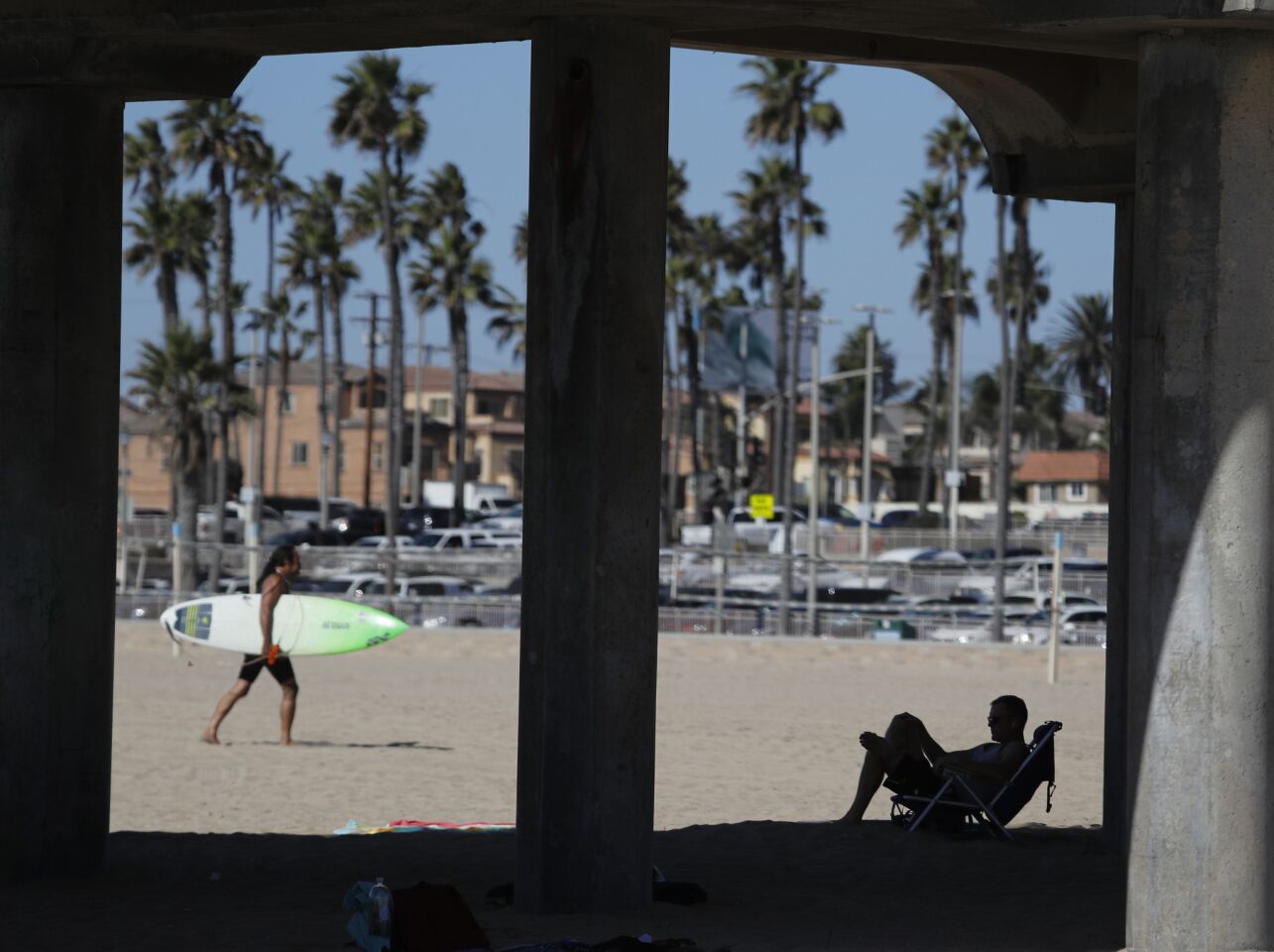 Jim Dodd of Huntington Beach relaxes in the shade under the Huntington Beach Pier as a surfer heads across the beach after riding large Santa Ana wind-swept waves amid 104-degree temperatures in Huntington Beach on Tuesday.