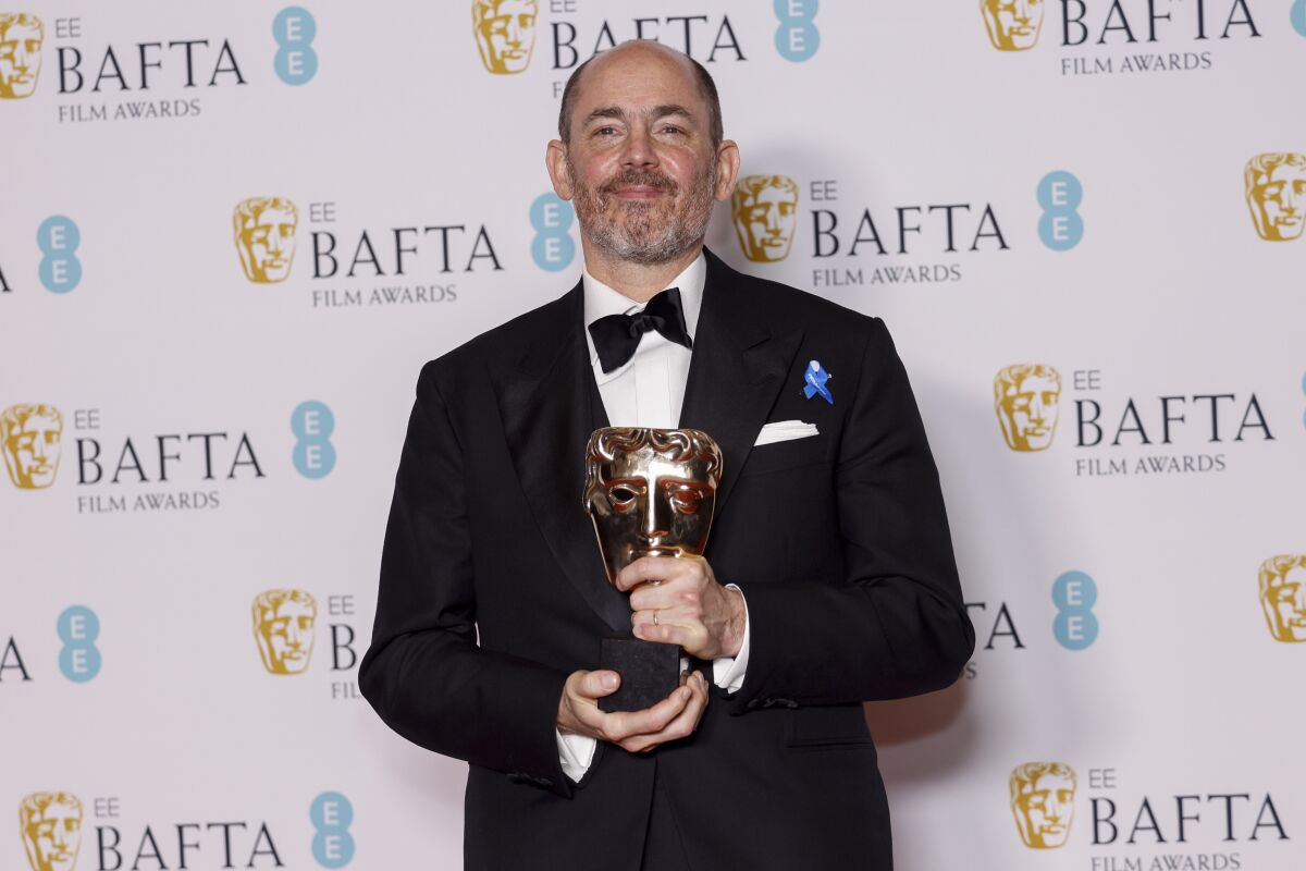 A man in a tuxedo holds an award in front of a BAFTA background