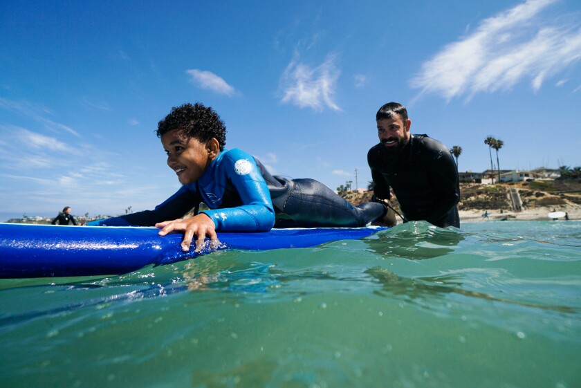 A man stands behind a child on a surf board in the ocean with the beach behind them