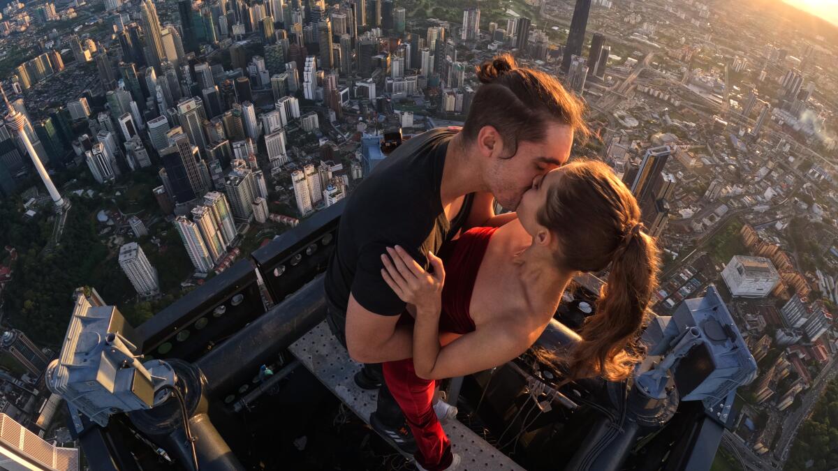 A couple embraces while perched on the rooftop of a building with the ground far below.