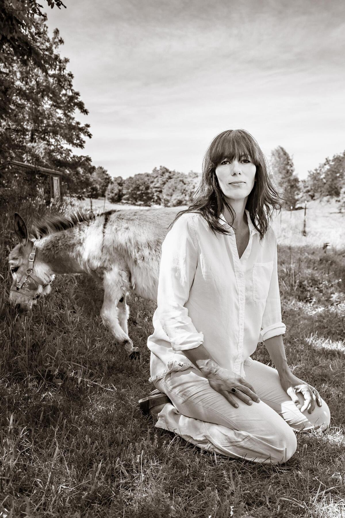 A woman in white sits in a field in front of a grazing animal.