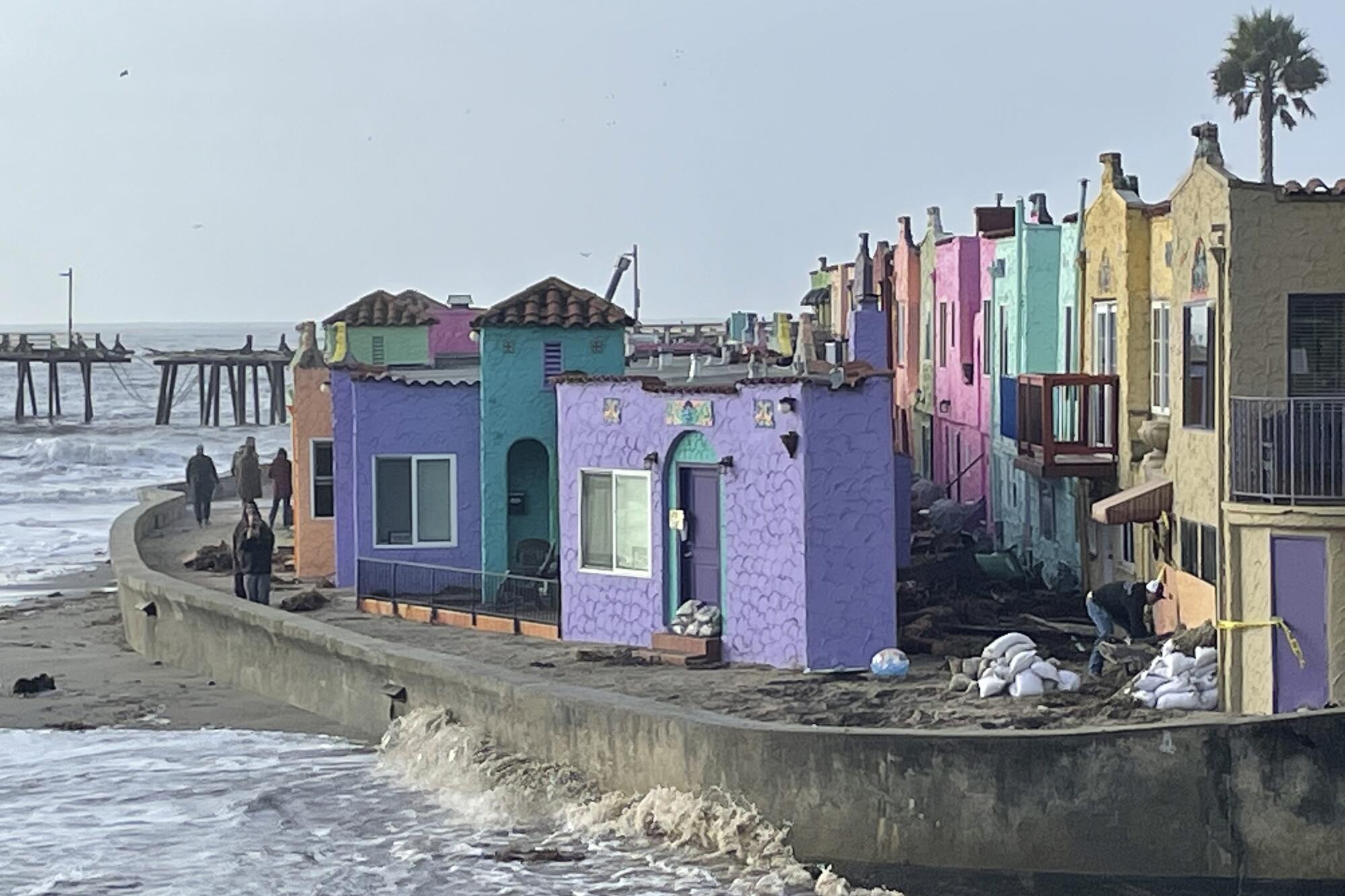 People walk through a storm-damaged section of a seaside grouping of buildings with facades of different pastel colors