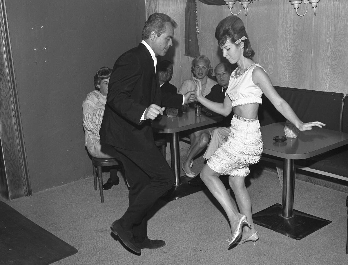 A man in a suit and a woman in a fringed skirt and beehive hairdo twist as they dance.