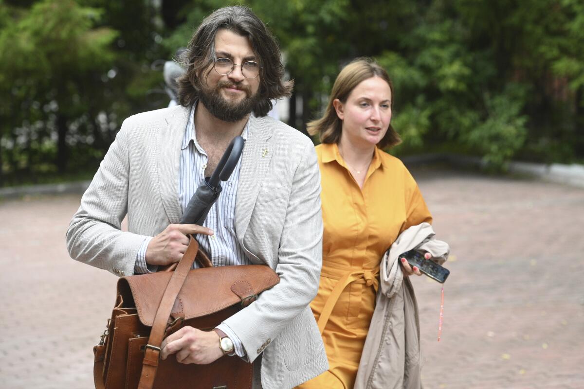 A man carrying a satchel and umbrella and a woman carrying a raincoat and phone walk together outdoors.