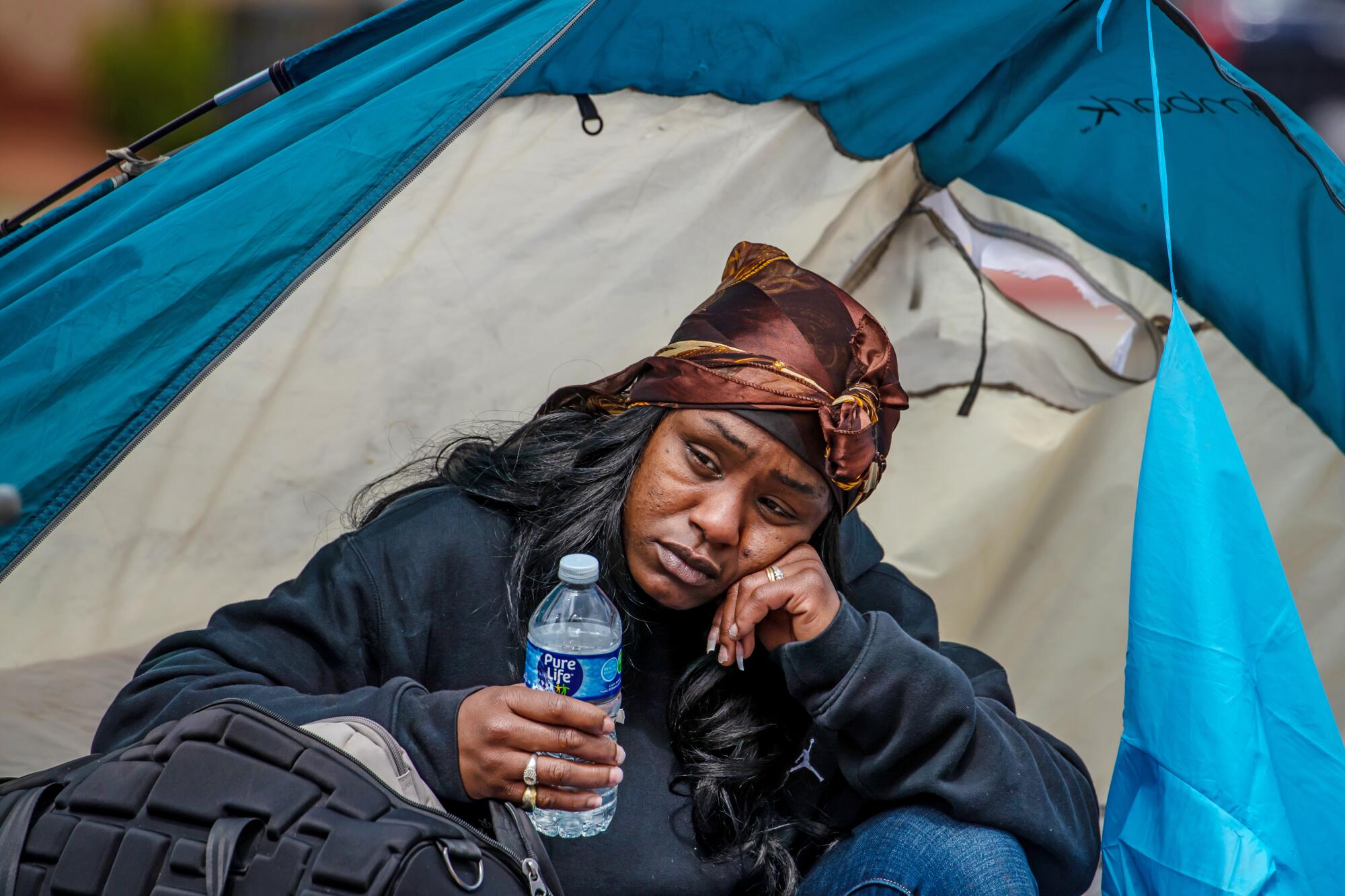 A head-and-shoulders frame of a woman sitting down in an opening of a tent, resting her head on her left hand, looking sad