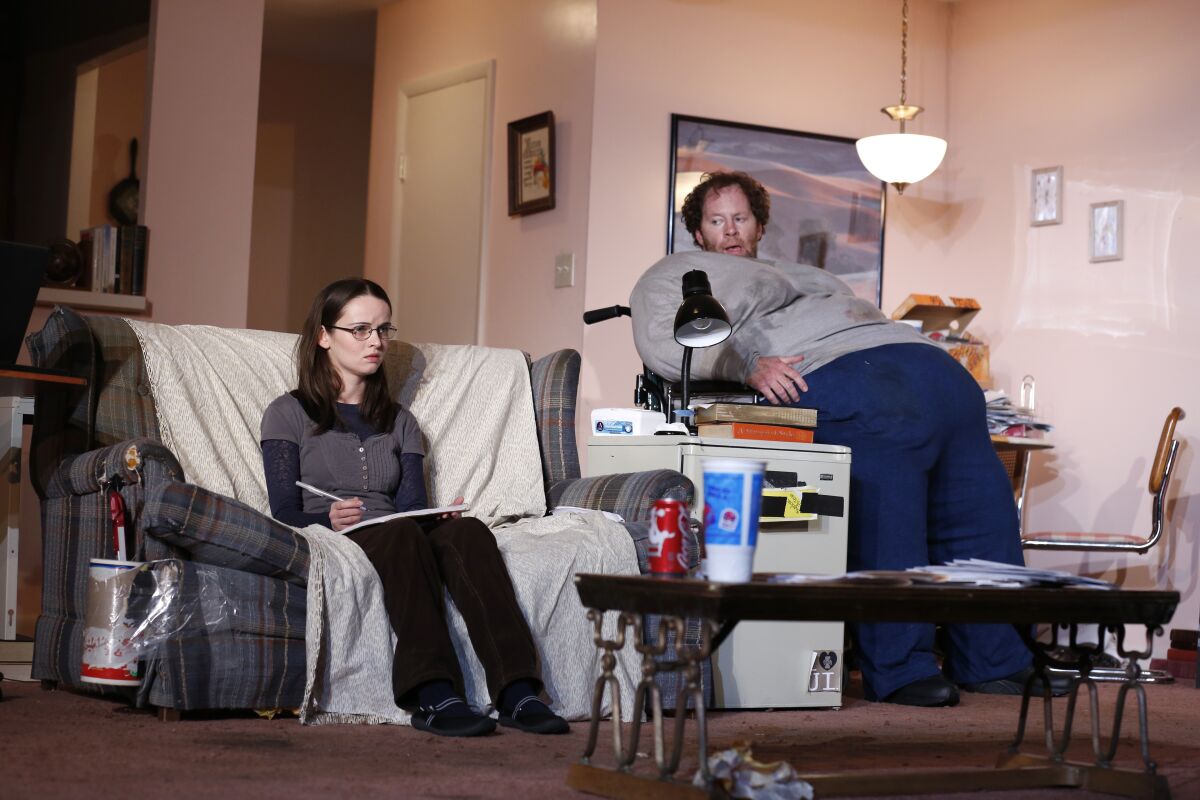 A young woman sitting on a couch near an obese man sitting on a desk in a play.