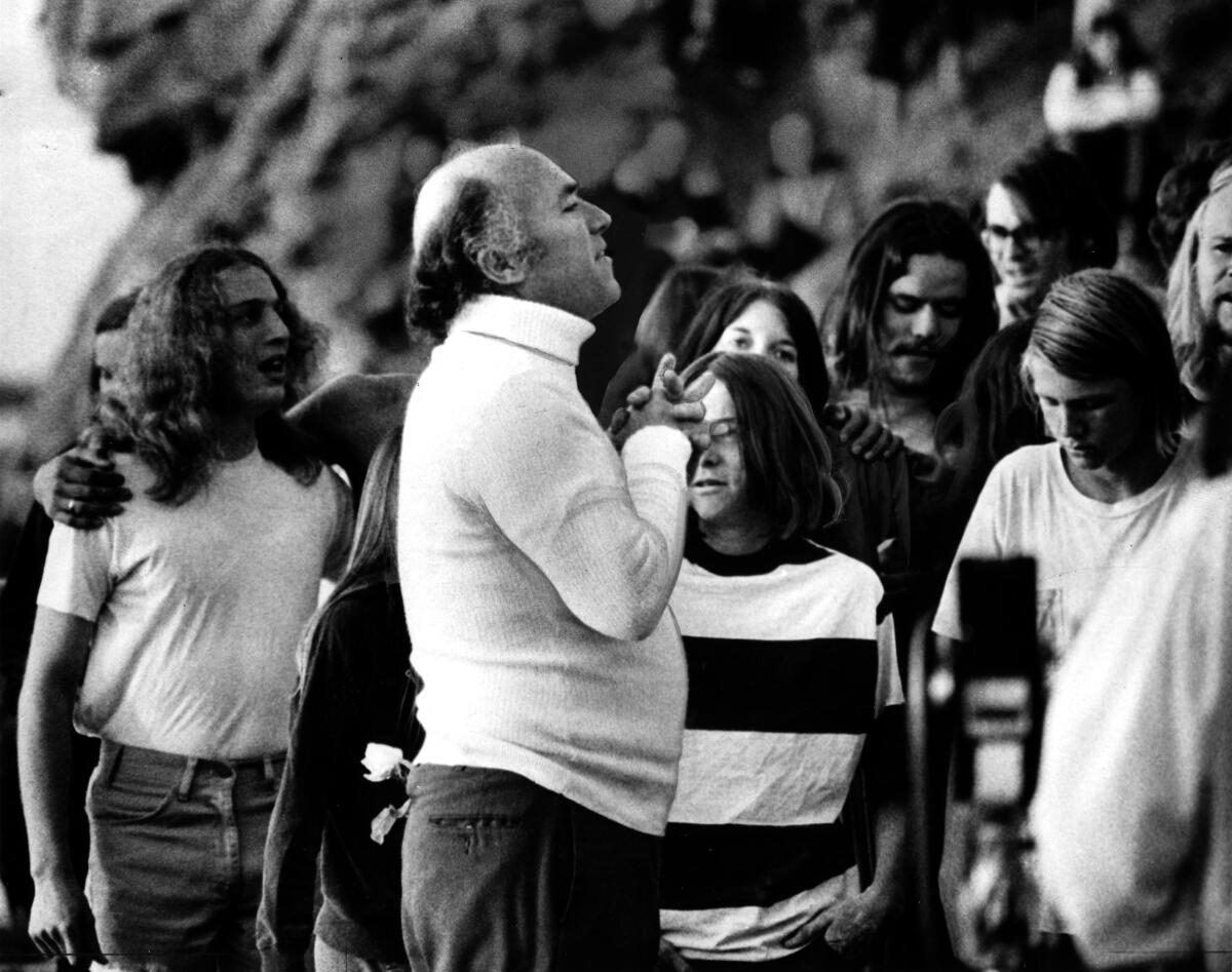 A man in a white turtleneck leads a group of young people in prayer.