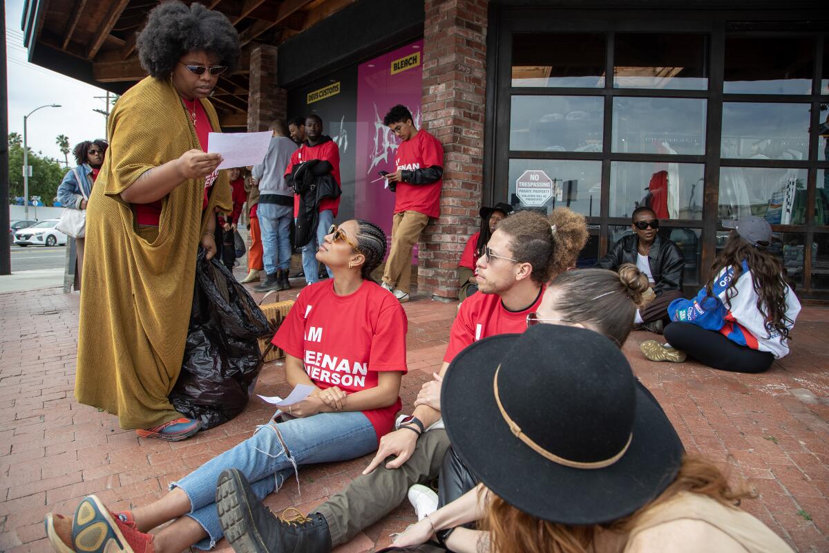 Patrisse Cullors stands lookimg at a document while other protesters sit on a sidewalk.