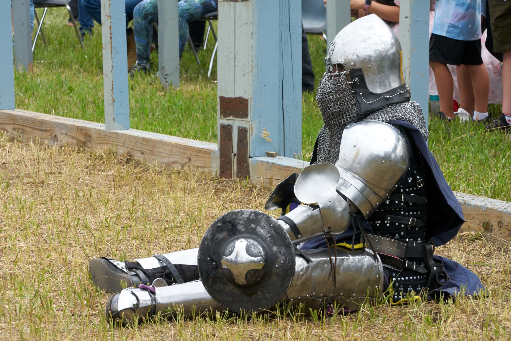 An armored tournament participant sits on the grass holding a shield. 