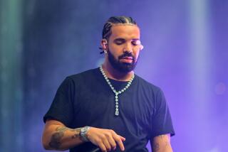 Man, Drake, hair in cornrows, on stage wearing black t shirt and diamond chain, holding mic along his chest, looking down 