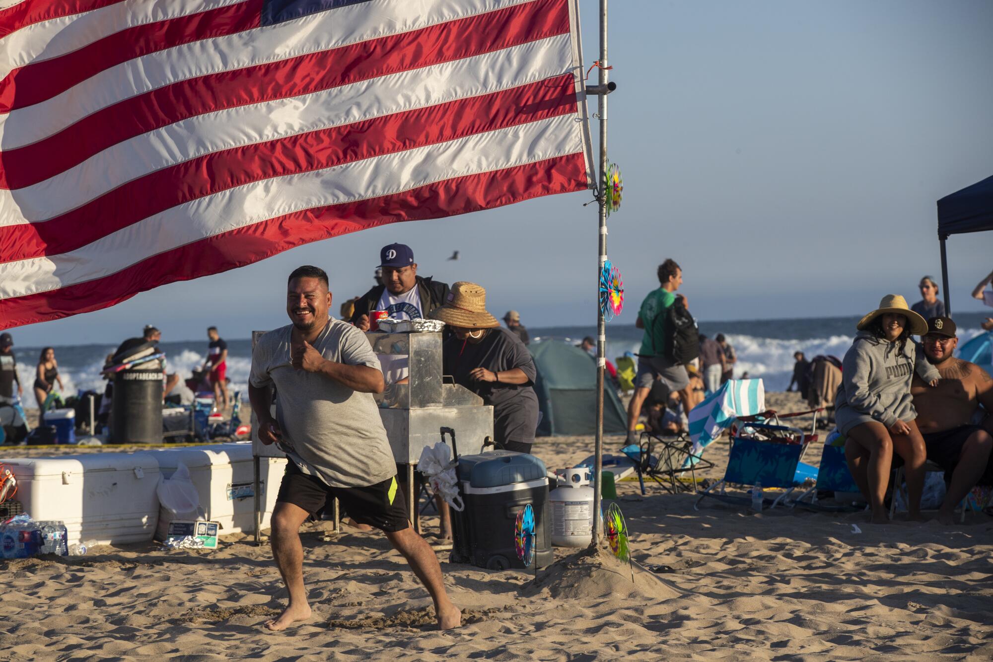 A smiling man stands on the sand with a U.S. flag and other people in the background 