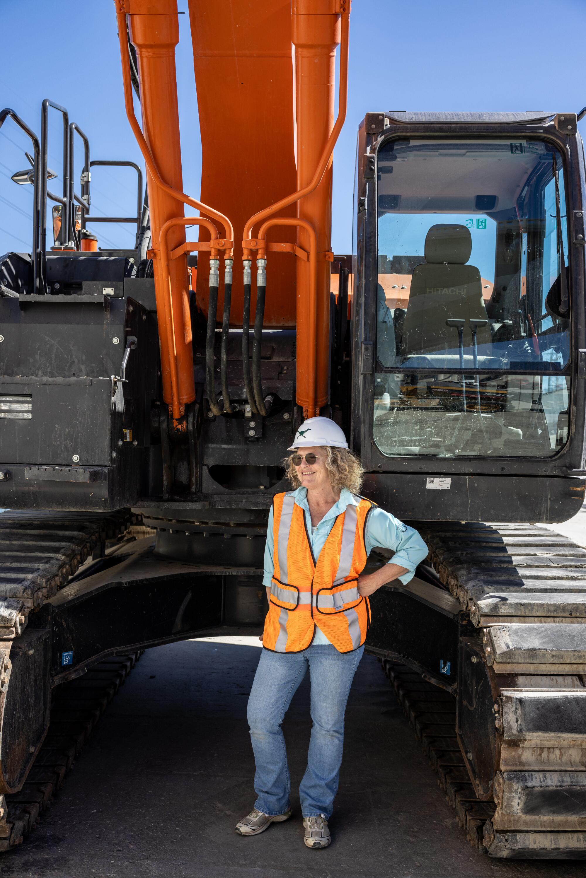 Artist Lauren Bon, in hard hat and a bright safety vest, stands next to an excavator in the concrete bed of the L.A. River