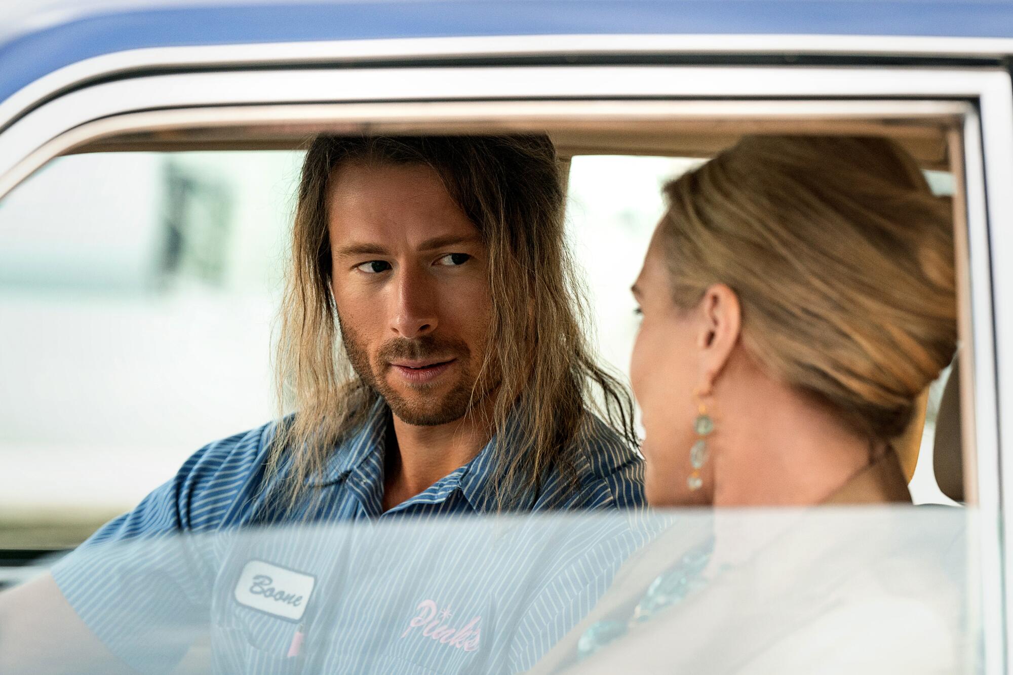 A man with scraggly long hair sits in a car talking to a woman