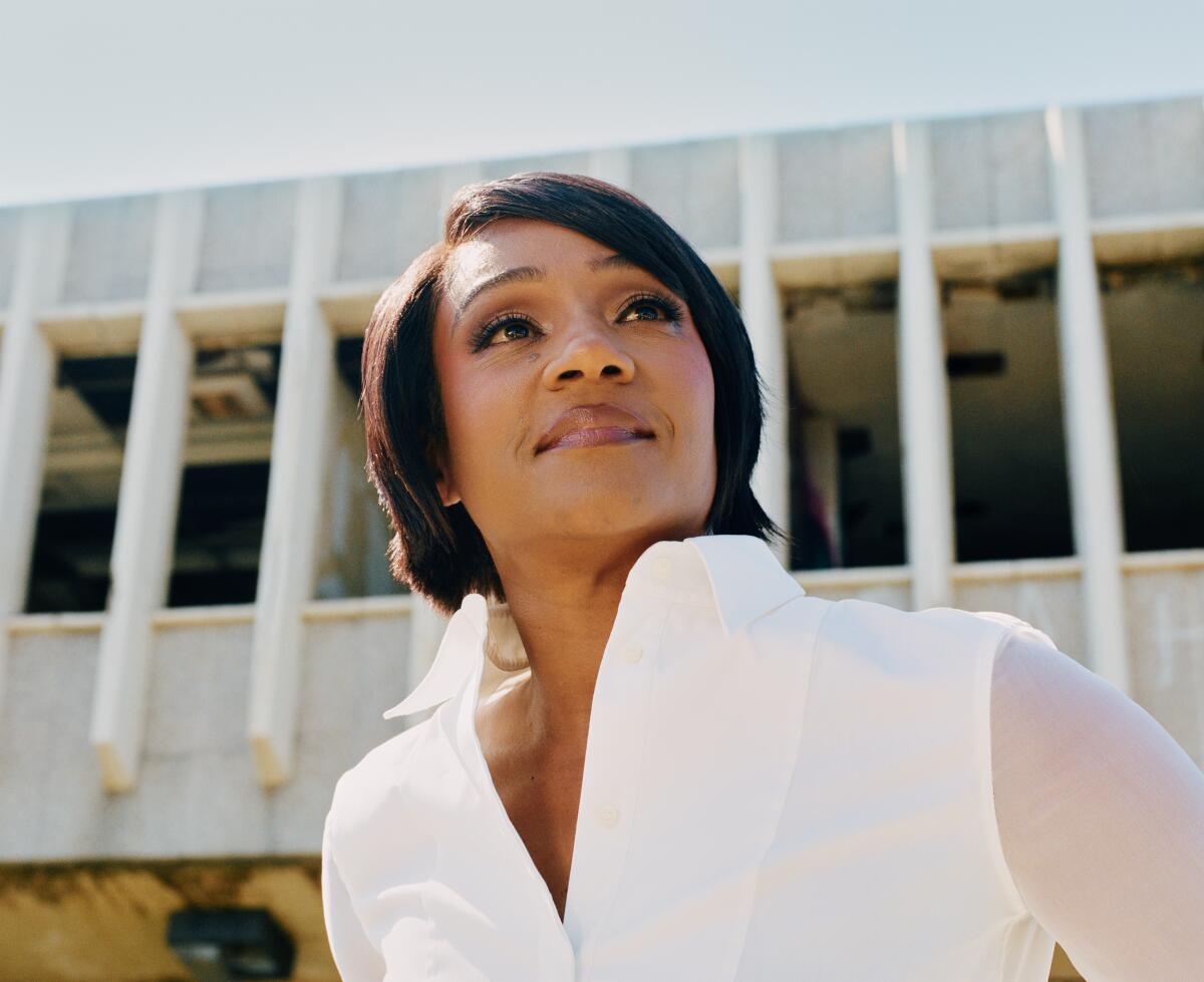 Tiffany Haddish looking up and to the side in a white collared blouse with sheer sleeves in front of a white building