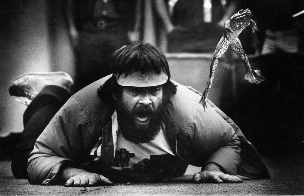 A bearded man lies on his stomach on the floor, yelling at a leaping frog.