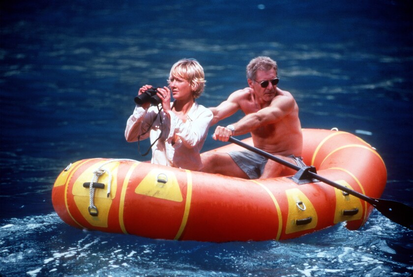 Anne Heche and Harrison Ford in an inflatable raft, her holding binoculars and him paddling.