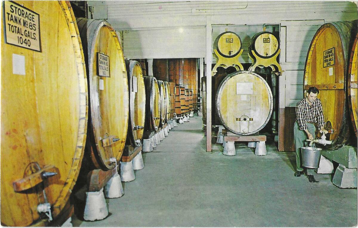 Large barrels of wine in a room where a man holds a bucket under a spigot.